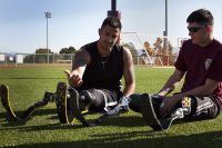 Two athletes with advanced running-blade prosthetic legs sitting together on a sports field