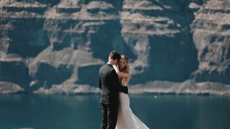 A bride and groom embracing on a cliff overlooking a lake and mountain cliffs