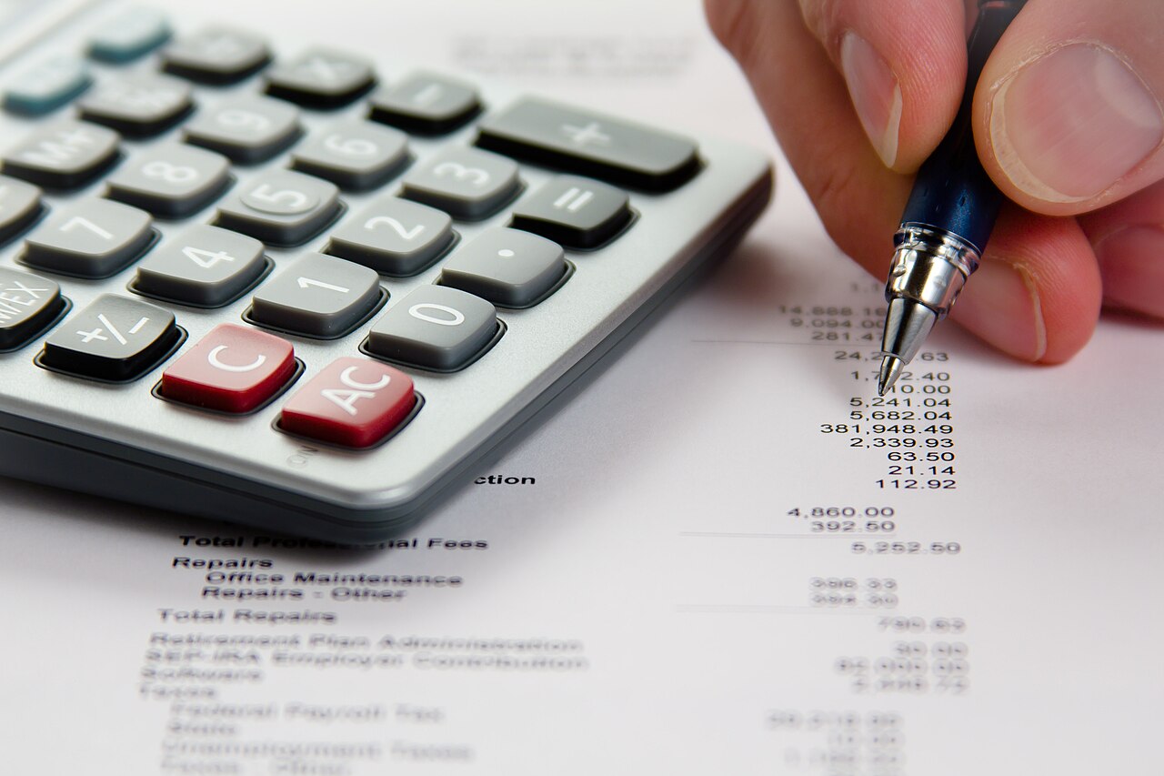 A hand with a pen and calculator reviewing a page of financial figures