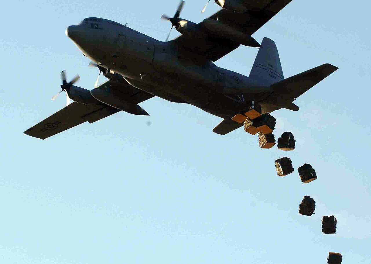 A cargo plane releasing a line of parachute bundles in mid-air over a field