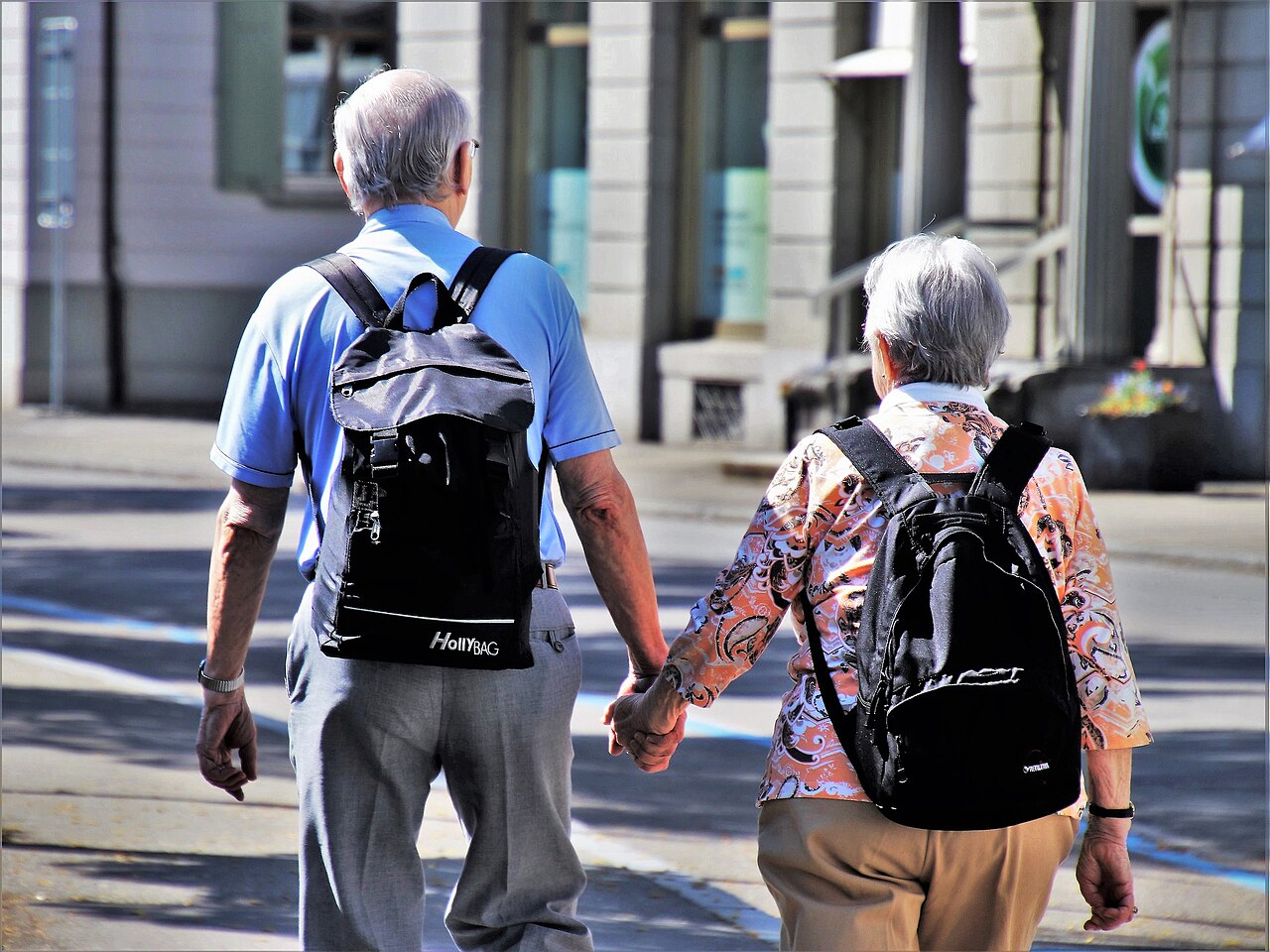 An elderly couple walking down a city street together, holding hands from behind