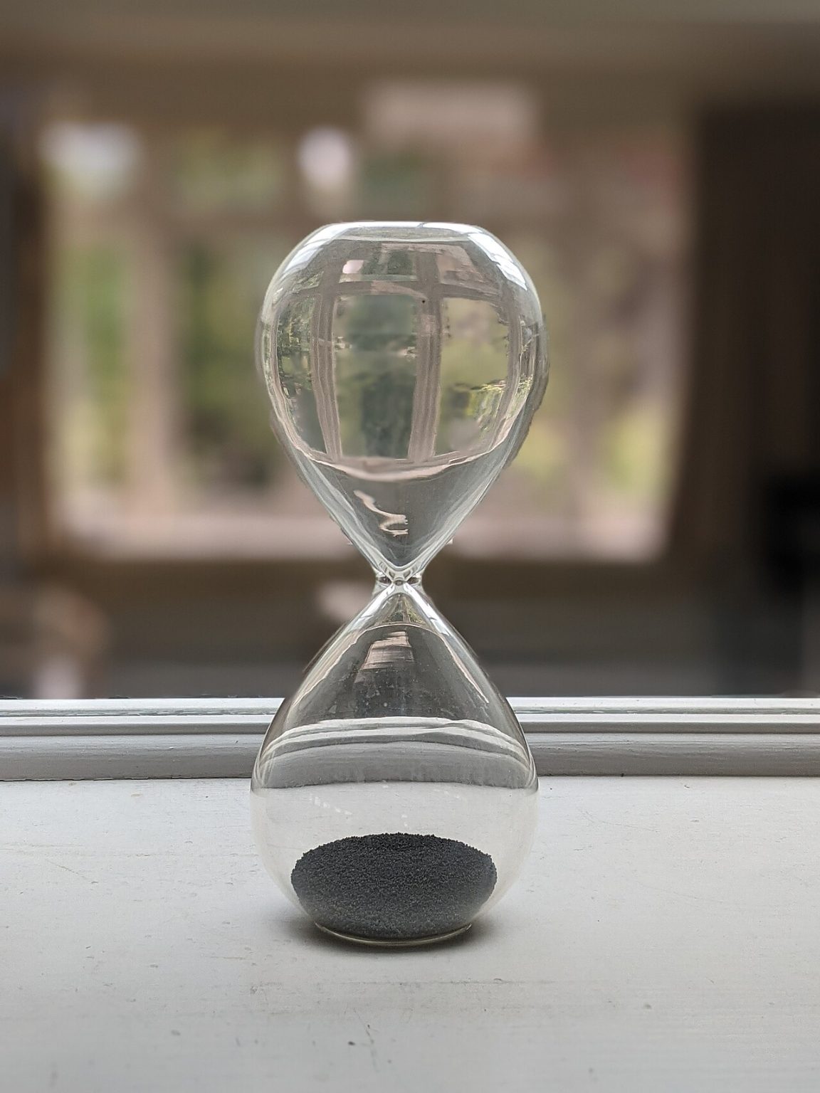 A clear glass hourglass with sand falling through it, sitting on a windowsill