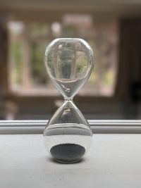 A clear glass hourglass with sand falling through it, sitting on a windowsill