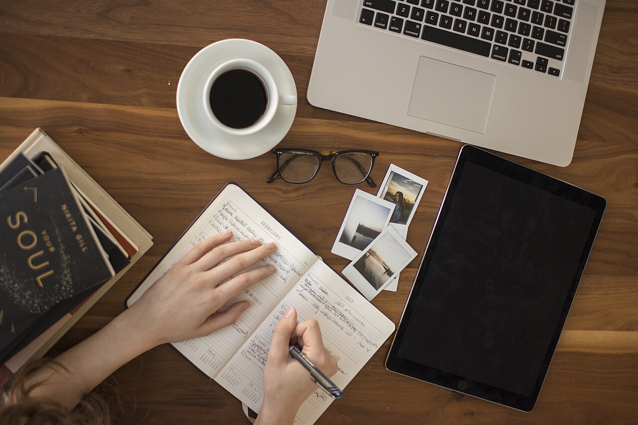 A person journaling in a notebook next to a cup of coffee and a laptop