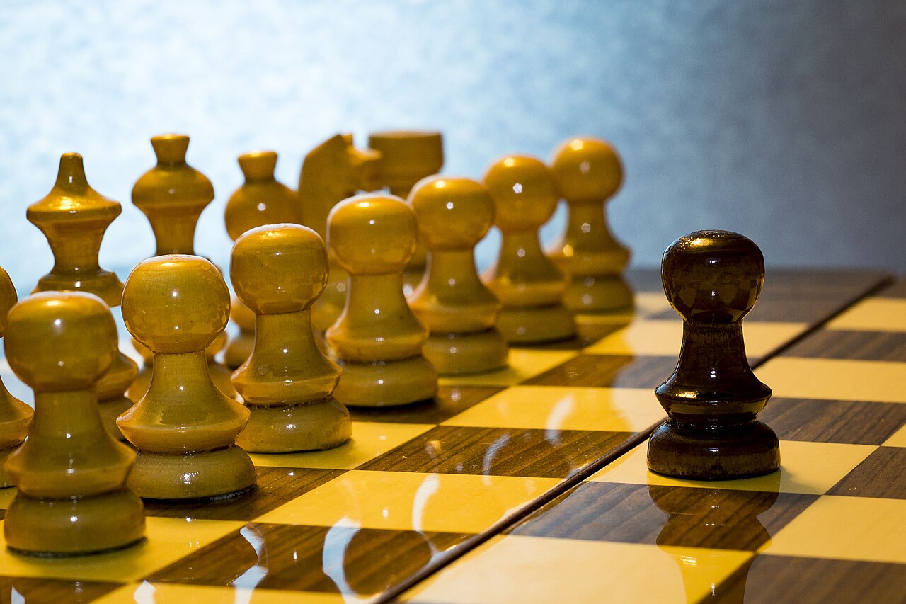 A single dark chess pawn standing alone opposite a full lineup of light-colored pieces on a board