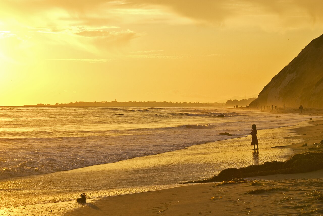 A lone silhouetted figure standing at the edge of the water on a beach at golden sunset