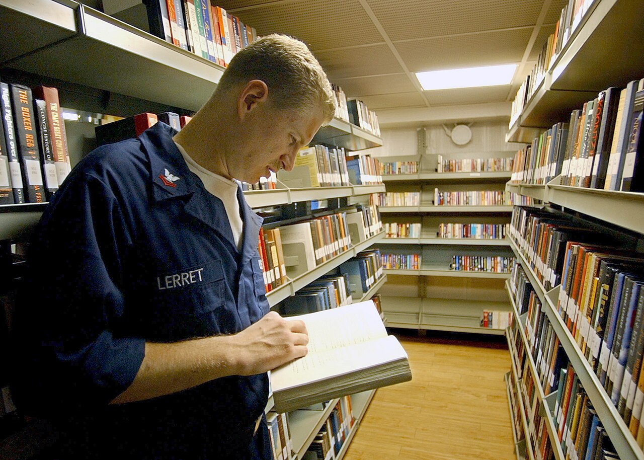A man reading a book while standing between tall library shelves