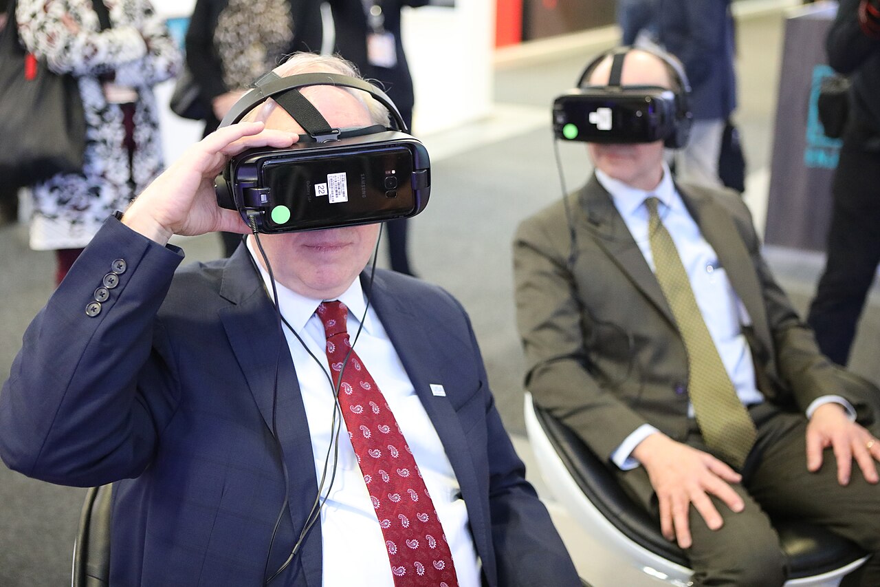 A man wearing a virtual reality headset with another person wearing one in the background