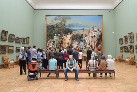 Museum visitors gathered in a gallery hall looking at a large classical painting