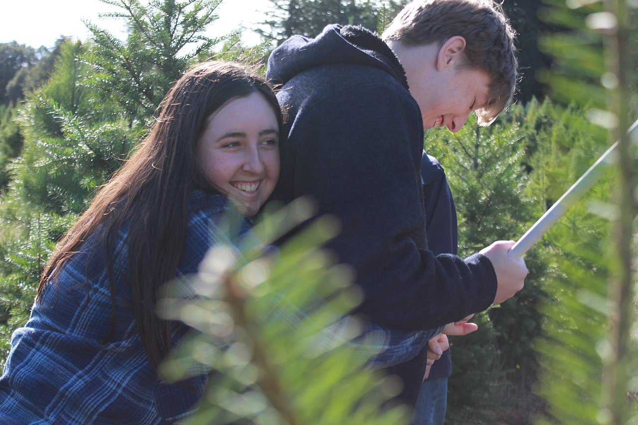 A young woman laughing while hugging her sibling from behind at an outdoor Christmas tree farm