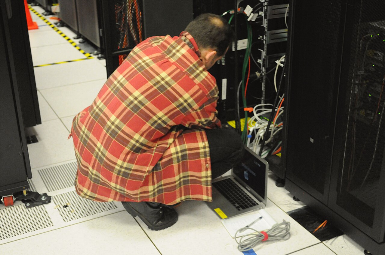 A technician crouching to work on server rack cabling in a data center, with a laptop on the floor
